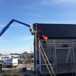 Montaż czarnego dachu panelowego na domu w trakcie budowy, pracownicy na podnośniku i rusztowaniu, widoczny ocieplony szkielet budynku i zimowy krajobraz w tle.