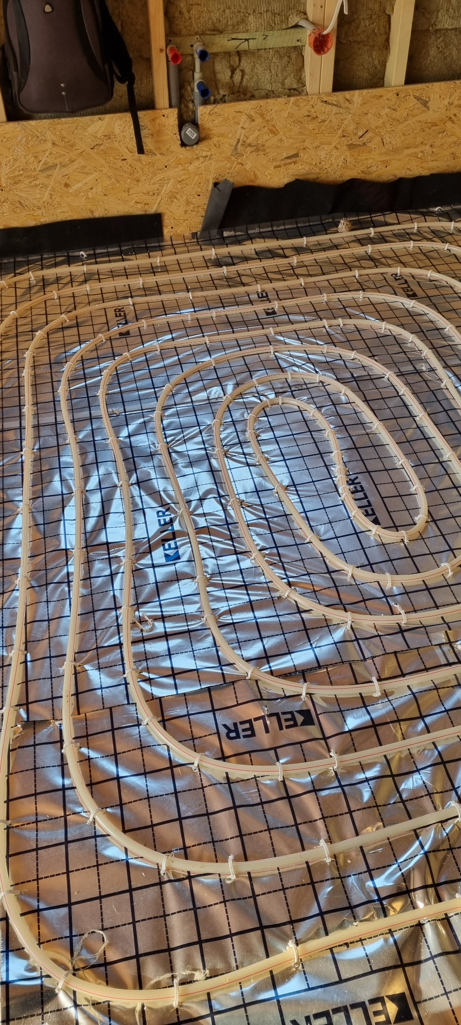 Rozłożona instalacja ogrzewania podłogowego z rurkami zamocowanymi do folii z nadrukowaną siatką, widoczne elementy instalacji wodnej na ścianie z płyt OSB w tle.