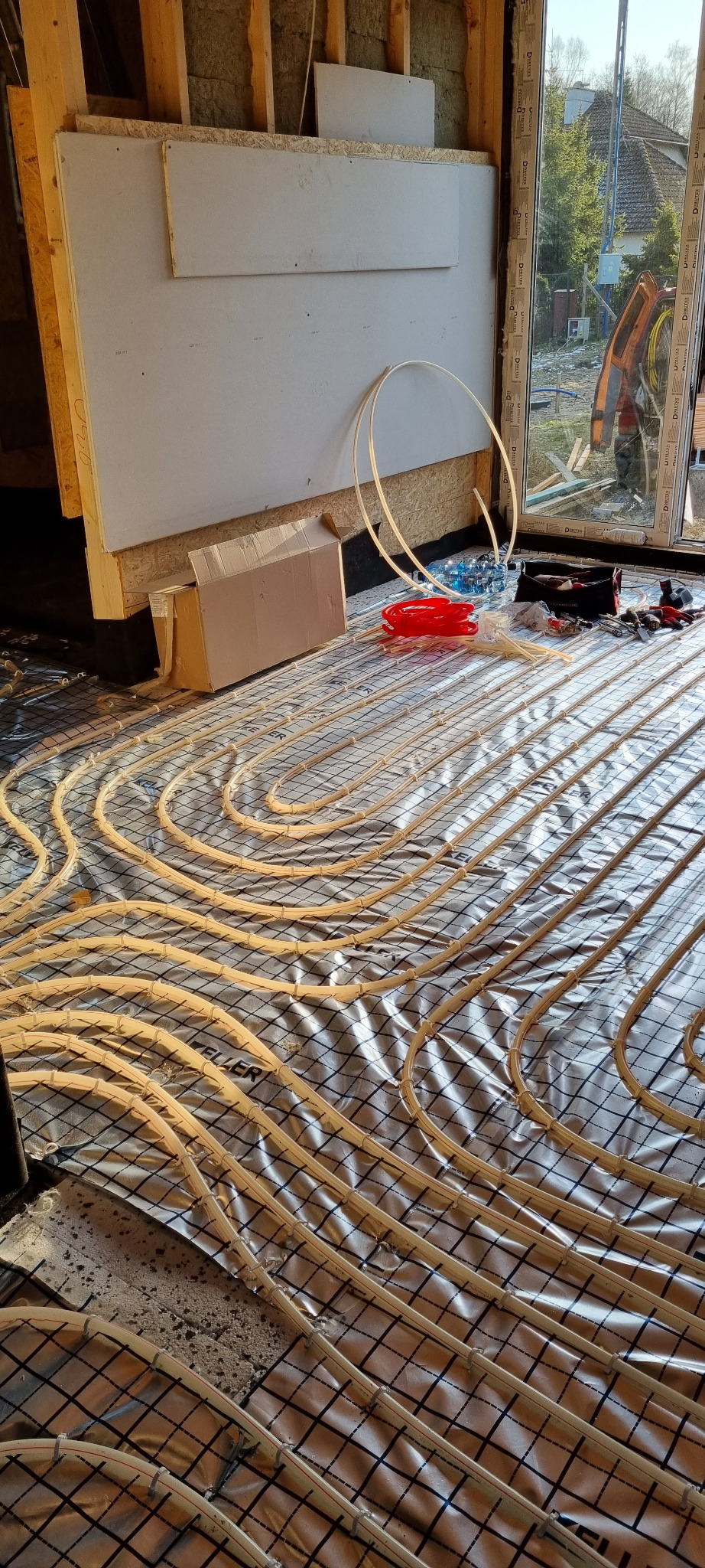 Rozłożone rury ogrzewania podłogowego na folii z siatką w trakcie montażu w nowym domu, widoczne materiały budowlane i narzędzia.