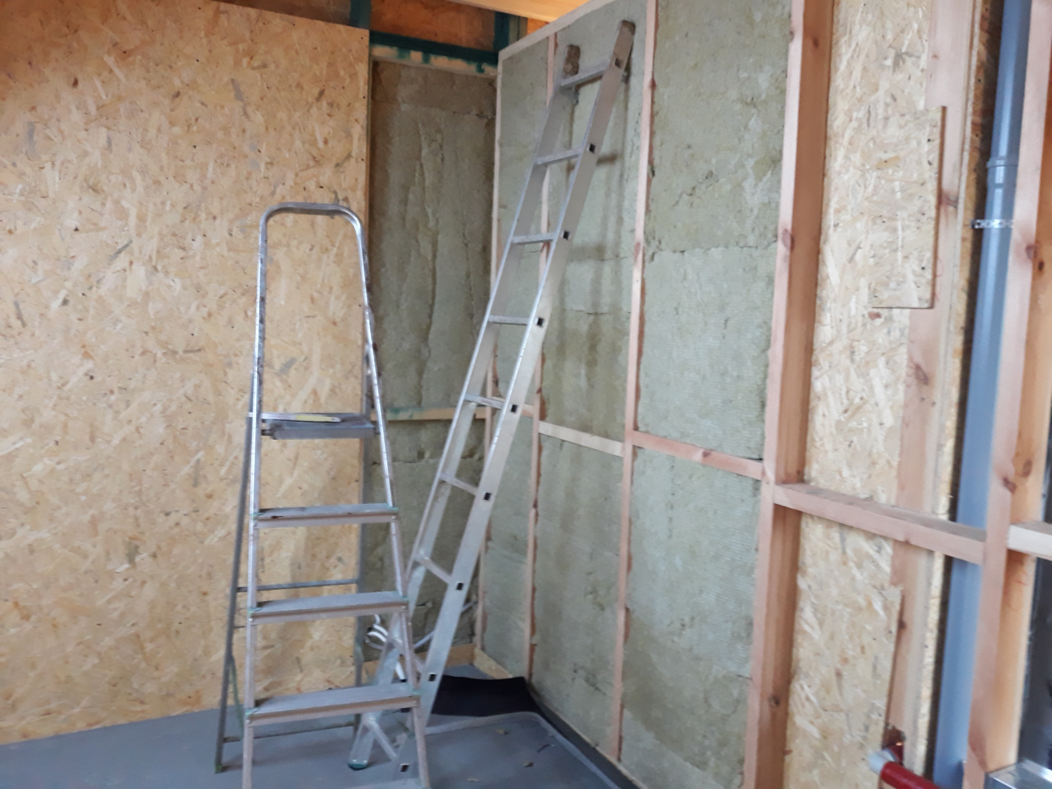 Wnętrze w trakcie budowy z widoczną konstrukcją ścian z drewna i płyt OSB, ocieplonych wełną mineralną. Dwie aluminiowe drabiny opierają się o ściany. Widoczna instalacja sanitarna.