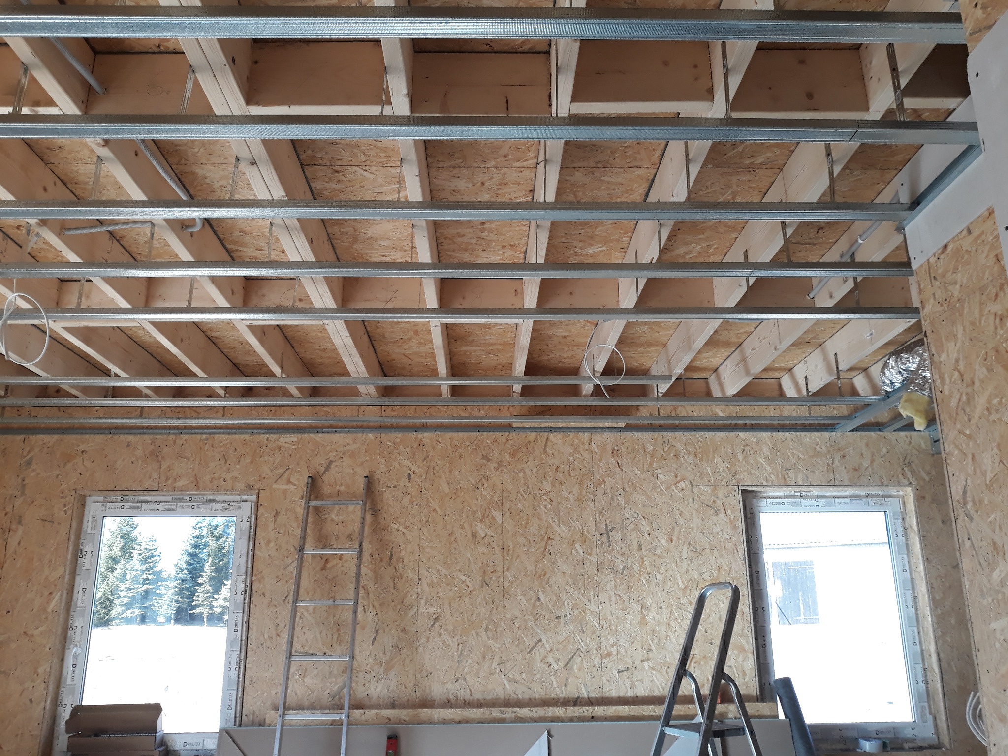 Wnętrze w trakcie budowy, widoczne drewniane elementy konstrukcyjne stropu, metalowe profile sufitowe, ściany z płyt OSB, okna z widokiem na zimowy krajobraz, dwie drabiny.