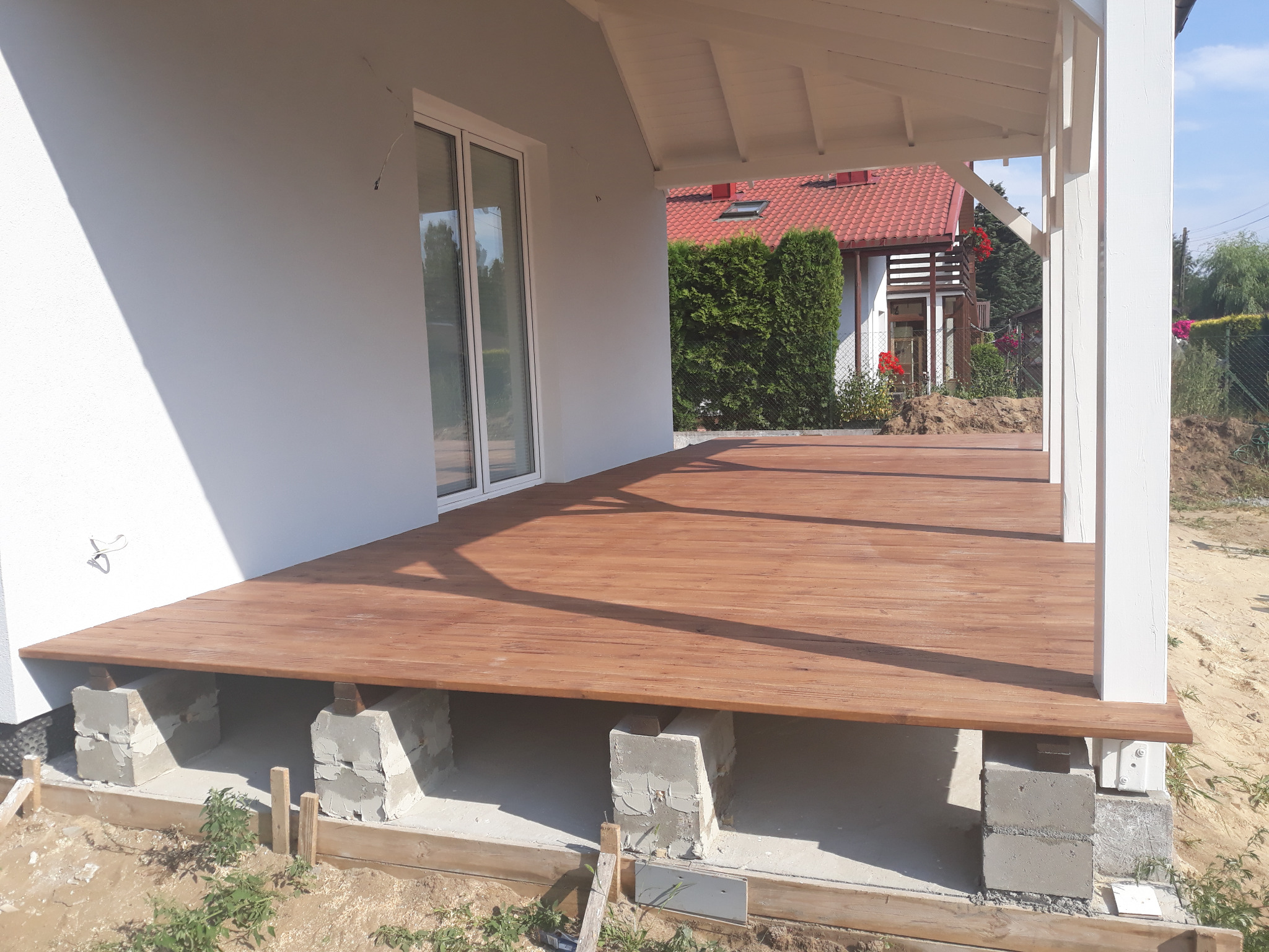 Drewniany taras przy białym domu w trakcie budowy, wsparty na betonowych bloczkach, widoczne fundamenty.