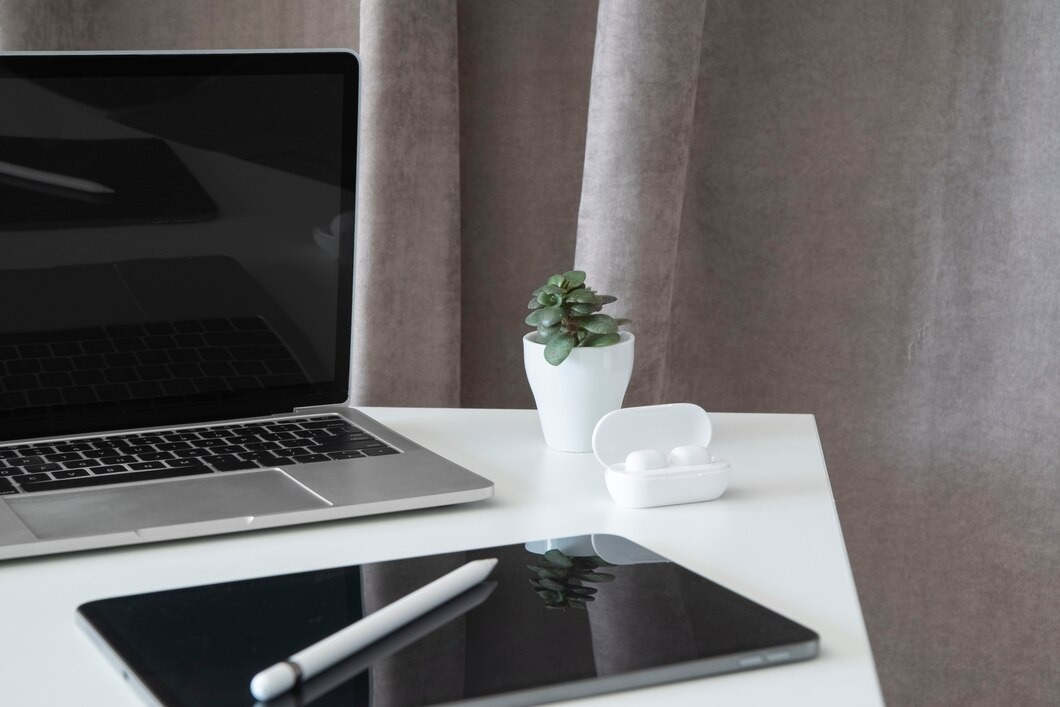 Laptop, tablet z rysikiem, słuchawki i roślina na białym biurku, na tle szarej zasłony. Minimalistyczne ujęcie sprzętu do edycji wideo.