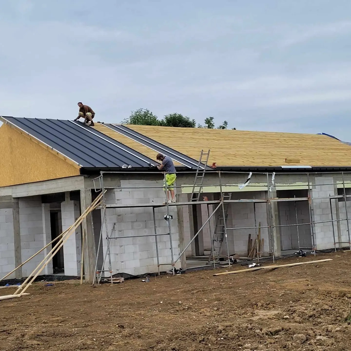 Dwóch pracowników na dachu w trakcie montażu pokrycia z blachy na nowym budynku z pustaków, jeden siedzi na dachu, drugi stoi na rusztowaniu.