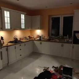 Jasna kuchnia z białymi szafkami z podświetleniem, widoczne niedokończone prace na podłodze, w tle okno.