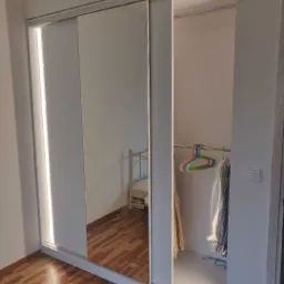 Biała szafa przesuwna z lustrzanymi drzwiami, oświetlenie LED po lewej stronie, widoczne ubrania wiszące w środku, parkiet na podłodze, odbicie pokoju w lustrze.