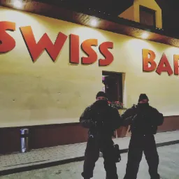 Dwóch umundurowanych mężczyzn z bronią stoi przed budynkiem z napisem 'SWISS BAR' w nocy, oświetlonym pomarańczowym światłem.