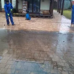Dwaj pracownicy w niebieskich kombinezonach czyszczą kostkę brukową za pomocą myjek ciśnieniowych, widoczna różnica w kolorze kostki przed i po umyciu, dom z drewnianymi elementami w tle.