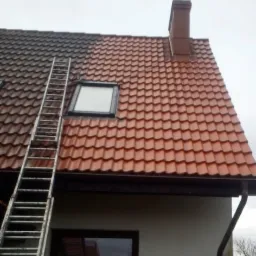 Częściowo umyty dach z czerwonej dachówki, widoczna drabina oparta o dach, okno dachowe i komin, dolna część elewacji budynku.