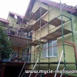 Dwupiętrowy dom w trakcie prac elewacyjnych, otoczony rusztowaniem, z widocznymi elementami dekoracyjnymi na narożnikach budynku i satelitą na dachu.