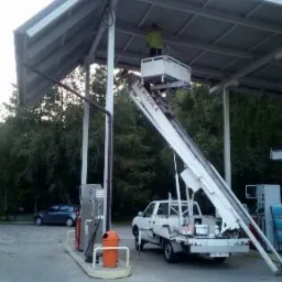 Pracownik w żółtej koszulce na podnośniku koszowym, wykonujący prace na zadaszeniu stacji benzynowej, widoczny dystrybutor i zaparkowany samochód.