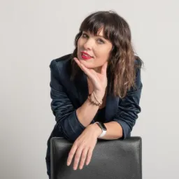 Portret kobiety w granatowej marynarce z aksamitu, opierającej się o czarne krzesło, z bransoletkami na nadgarstku i zegarkiem na ręce, uśmiechająca się z ręką pod brodą.