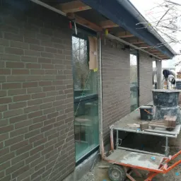 Murowanie ściany z cegły klinkierowej, widoczna świeżo wymurowana ściana z oknami, rusztowanie robocze z wiadrami i zaprawą, sznur murarski naciągnięty wzdłuż ściany, przygotowanie do dalszych prac.