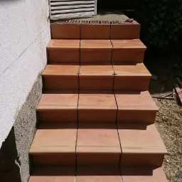 Kaskadowe schody zewnętrzne wyłożone terakotą z delikatnym zdobieniem, widoczne narzędzia glazurnicze i wiadro z zaprawą w tle.