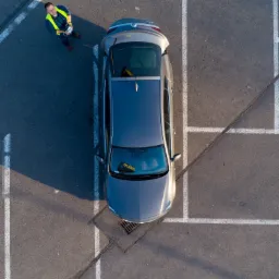 Zdjęcie z drona przedstawia mężczyznę w kamizelce odblaskowej, który kontroluje drona stojąc obok zaparkowanego samochodu na parkingu z wyznaczonymi miejscami postojowymi.