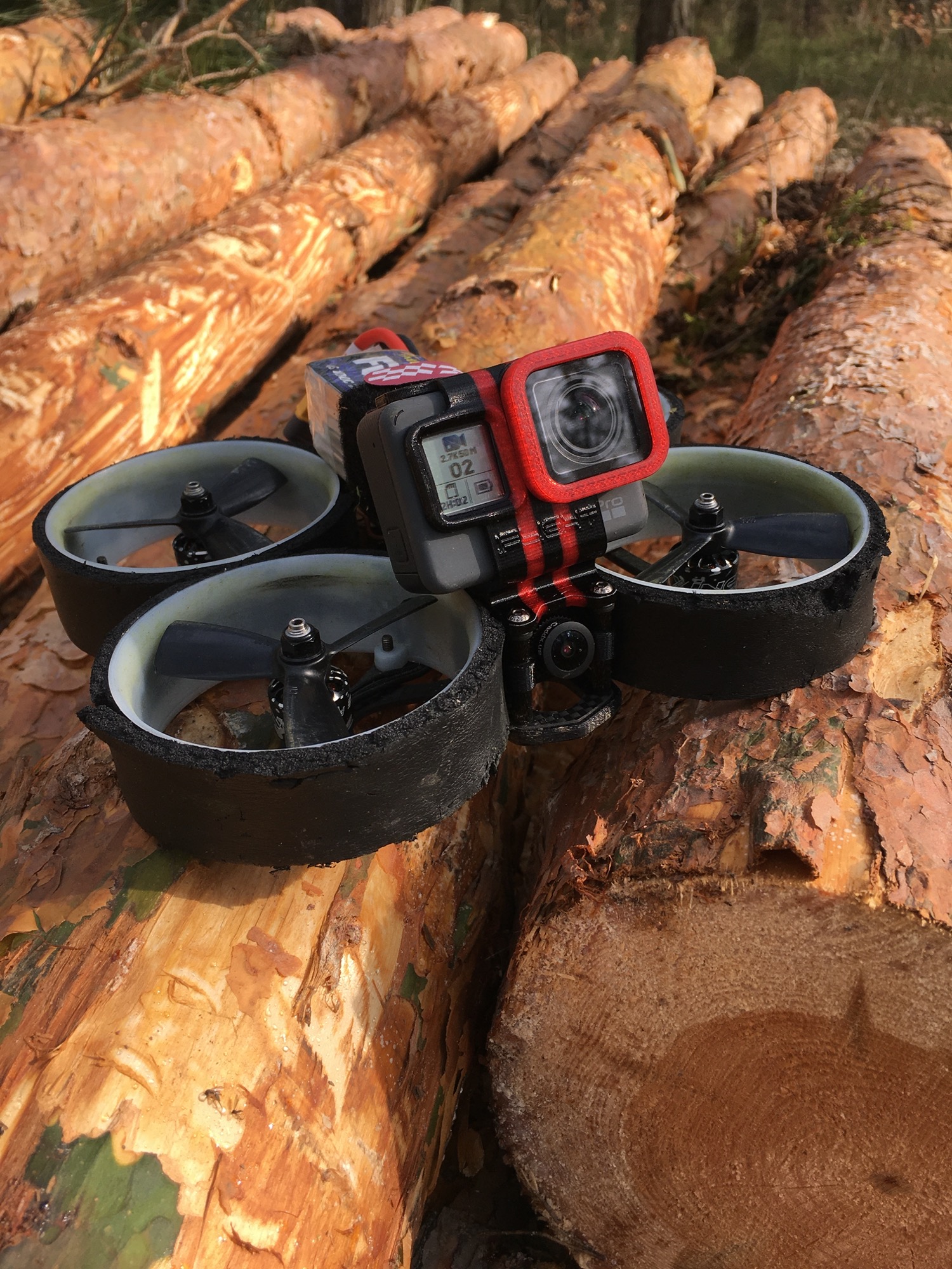 Niewielki dron wyścigowy z zamontowaną kamerą sportową, leżący na stosie świeżo ściętych pni drzew iglastych w lesie.