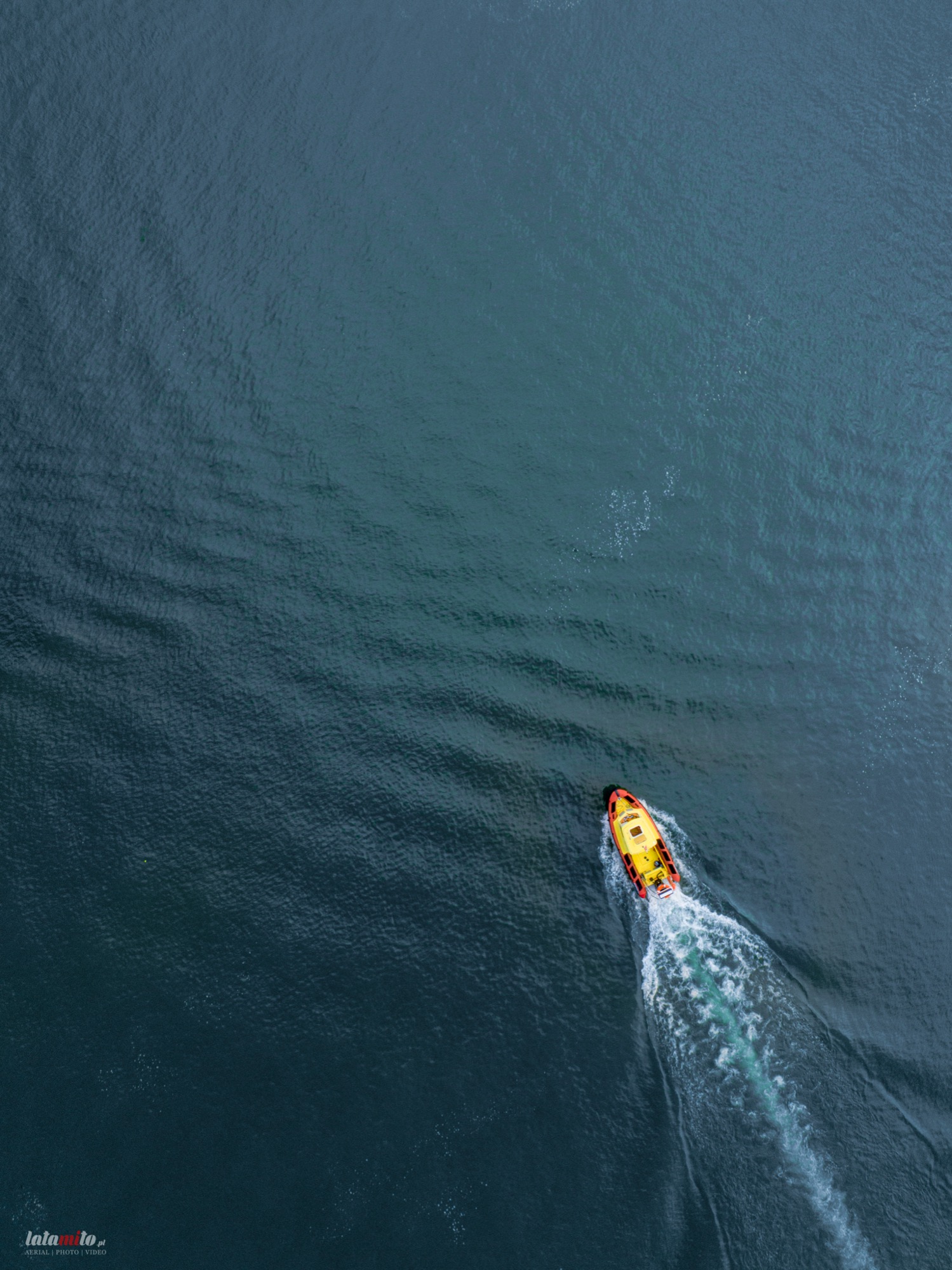 Żółto-czerwona motorówka płynie po ciemnej wodzie, widok z góry, tworząc biały ślad na powierzchni.