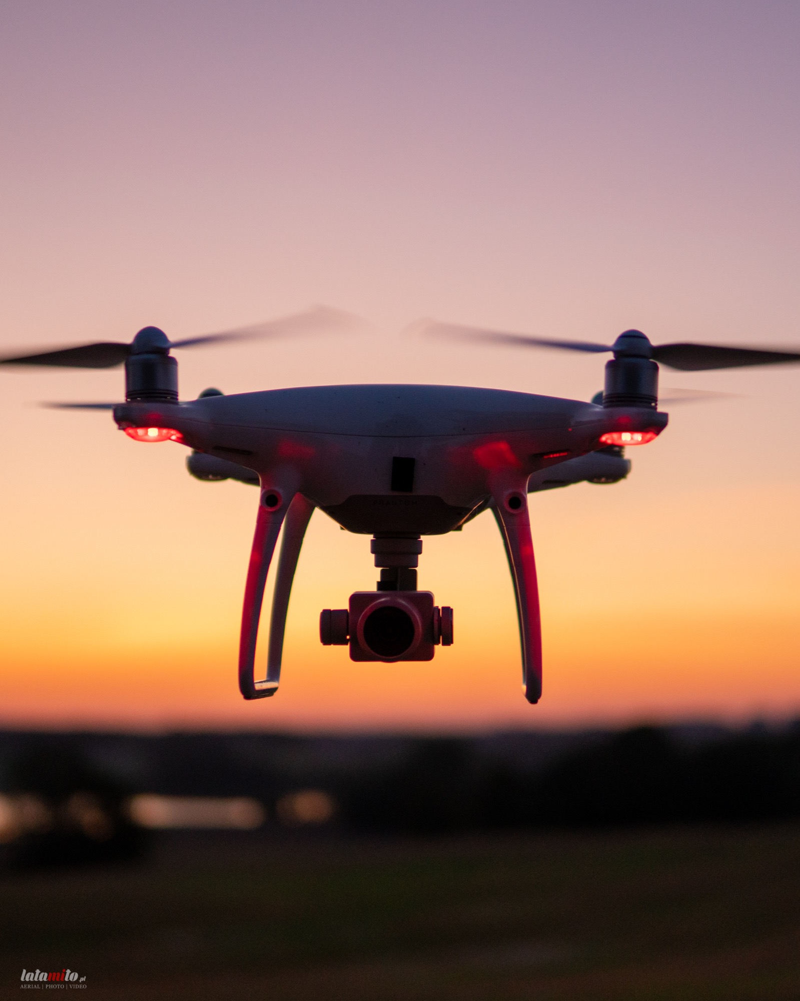 Latający dron z kamerą na tle zachodzącego słońca, z włączonymi czerwonymi lampkami pozycyjnymi.