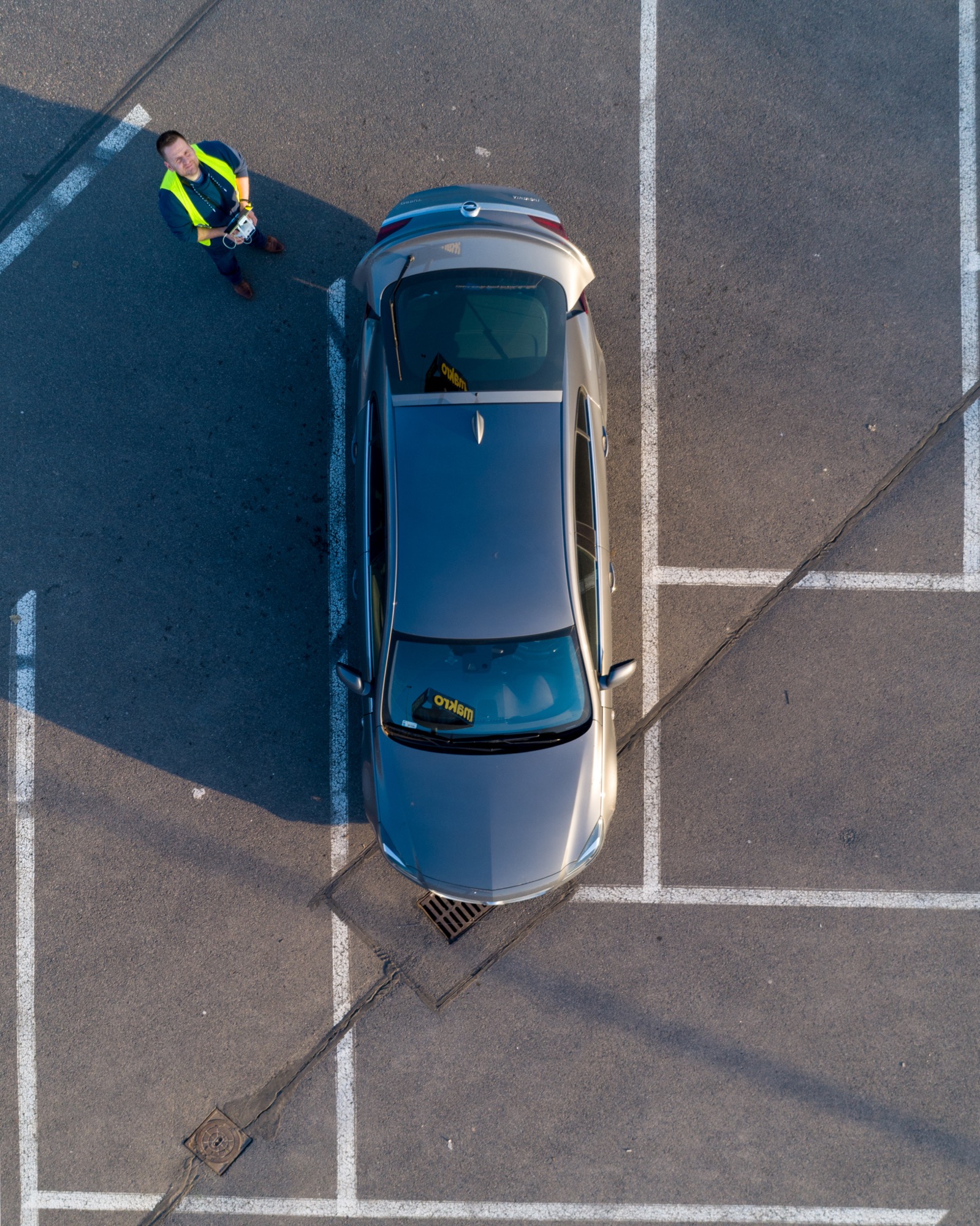 Zdjęcie z drona przedstawia mężczyznę w kamizelce odblaskowej, który kontroluje drona stojąc obok zaparkowanego samochodu na parkingu z wyznaczonymi miejscami postojowymi.
