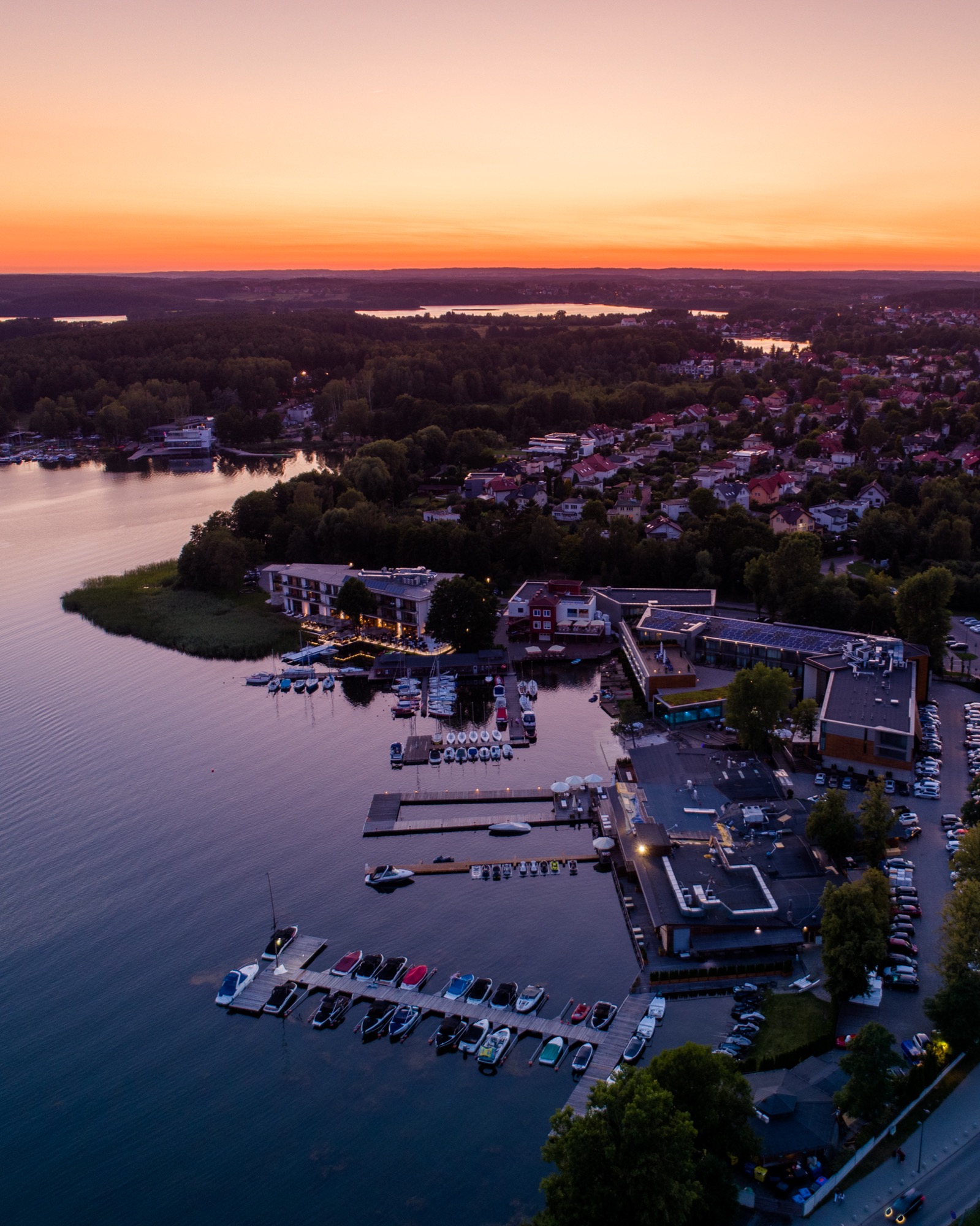 Panoramiczny widok z lotu ptaka na przystań jachtową o zmierzchu, z pomarańczowym niebem nad lasem i jeziorem, gdzie cumują łodzie przy drewnianych pomostach, a w tle widoczna zabudowa miejska...