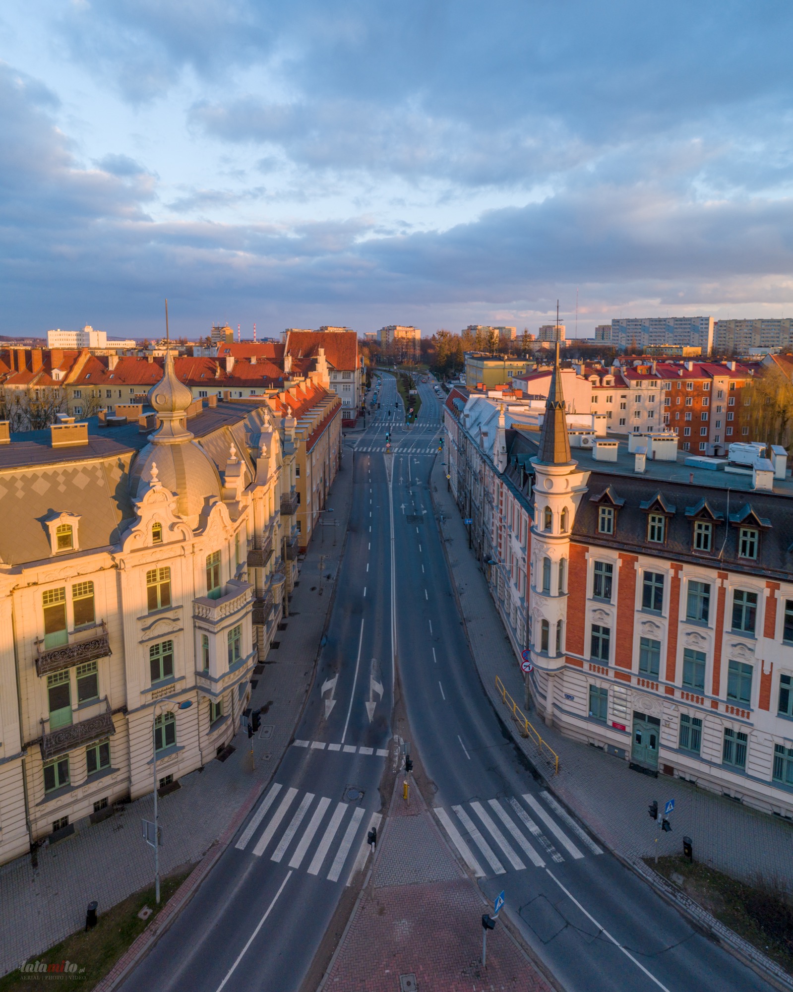 Panoramiczny widok z lotu ptaka na pustą ulicę miejską z zabytkową architekturą po obu stronach, wczesnym rankiem, z lekkim oświetleniem słonecznym i pochmurnym niebem.