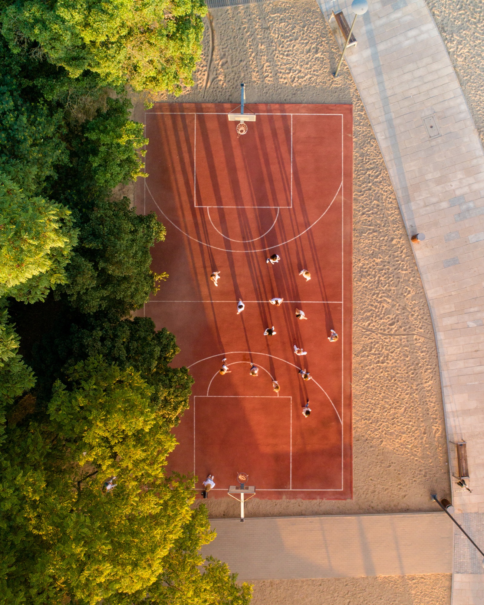 Widok z lotu ptaka na czerwone boisko do koszykówki otoczone zielenią i piaskiem, na którym grają ludzie, rzucając długie cienie w popołudniowym słońcu.