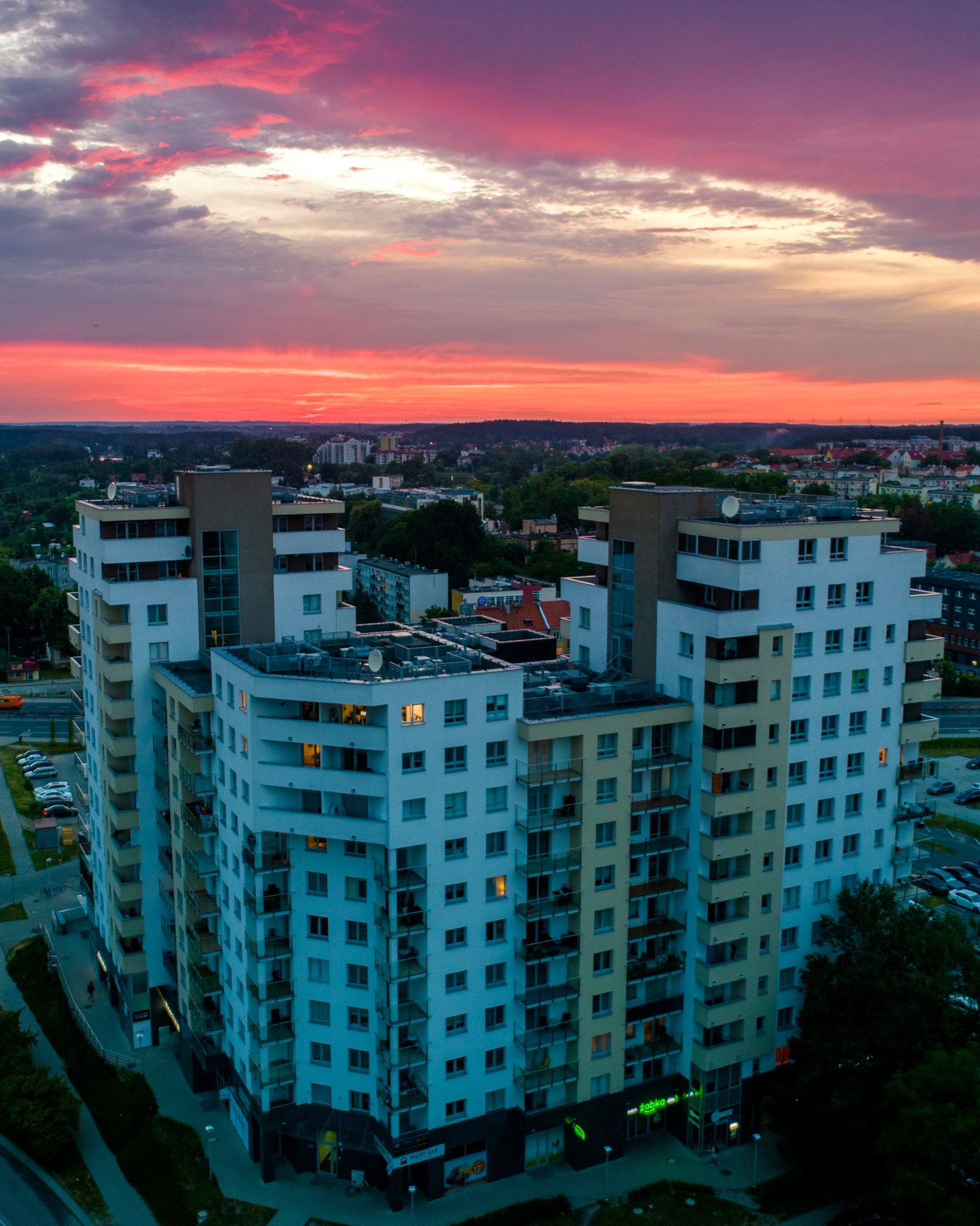 Wysoki budynek mieszkalny z balkonami, sfotografowany z góry o zmierzchu, z intensywnym czerwono-fioletowym niebem i widocznymi światłami w niektórych oknach.