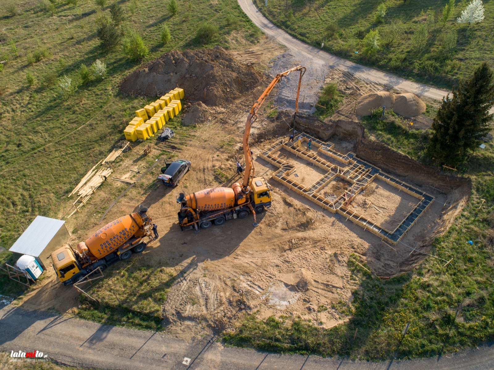 Widok z lotu ptaka na plac budowy fundamentów domu jednorodzinnego z dwoma betoniarkami, pompą do betonu, szalunkiem i materiałami budowlanymi.