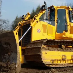 Żółty spychacz marki Dressta z nabraną ziemią, widoczny operator w kabinie, na tle drzew.