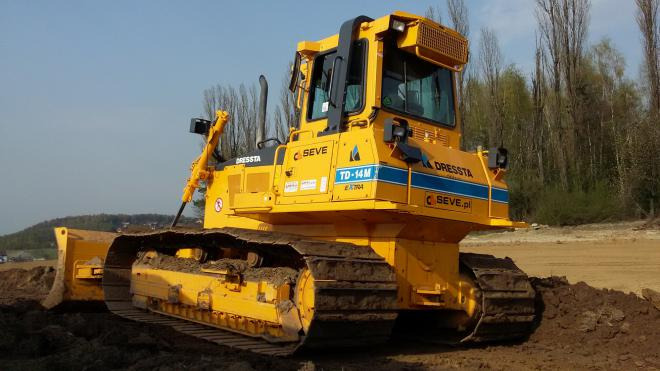 Żółty spychacz marki Dresssta TD-14M EXTRA na placu budowy, z widocznymi gąsienicami i lemieszem zagłębiającym się w ziemię, z lasem w tle.