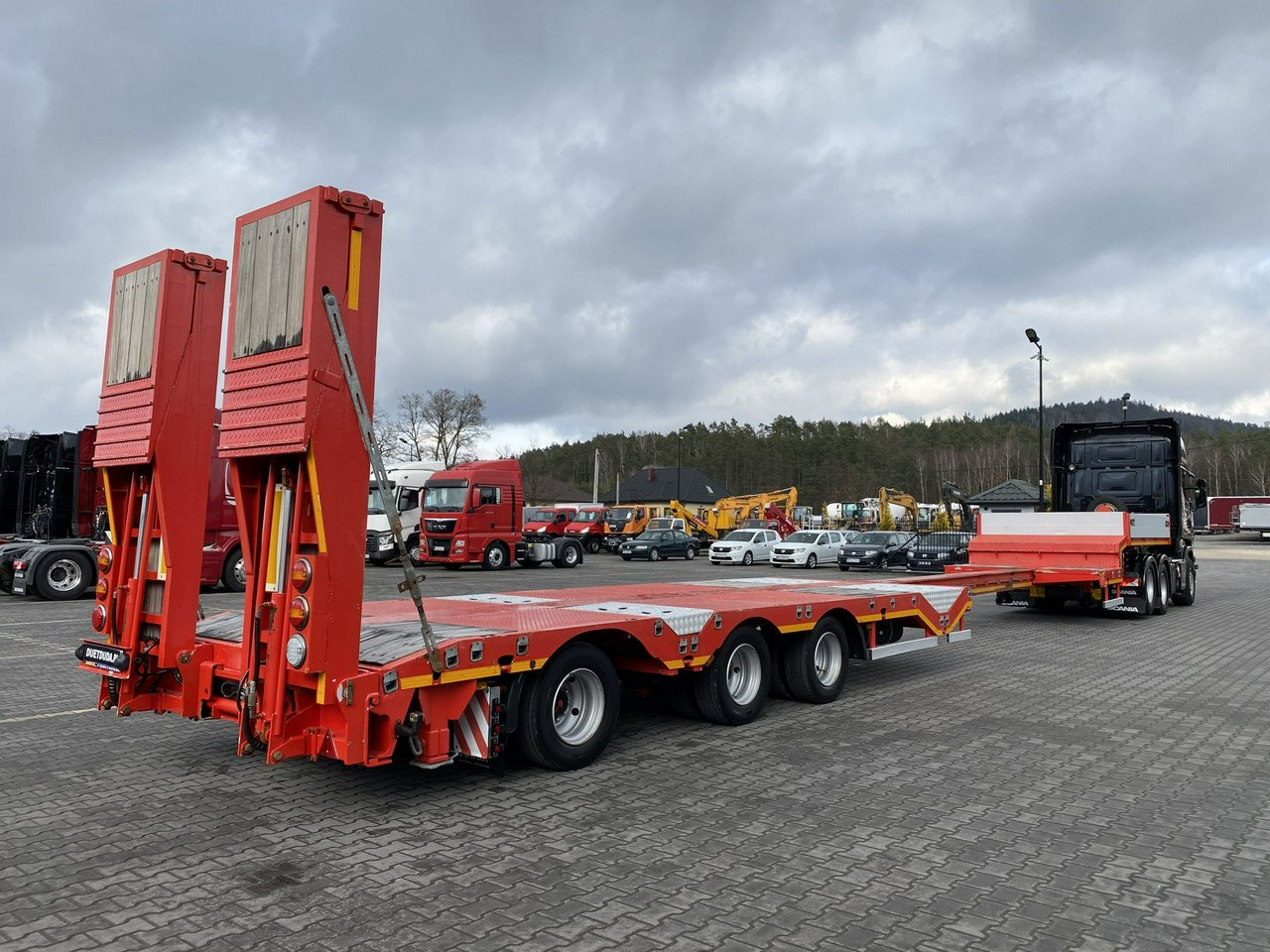 Czerwona naczepa niskopodwoziowa z podnoszonymi rampami załadowczymi, zaparkowana na placu z innymi pojazdami ciężarowymi i maszynami budowlanymi w tle, pochmurne niebo.