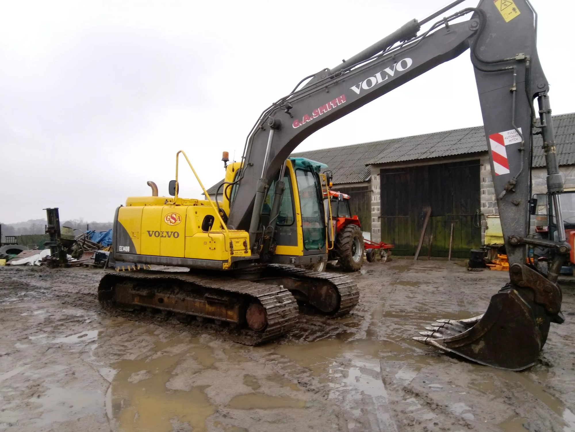 Żółto-szara koparka Volvo EC140B stojąca na błotnistym terenie, z widocznym budynkiem gospodarczym w tle i traktorem obok.