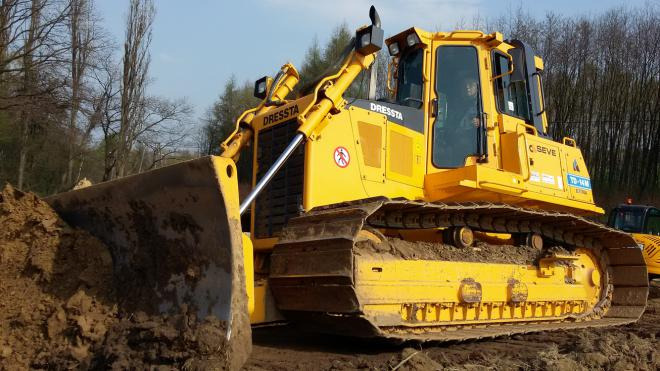 Żółty spychacz marki Dressta z nabraną ziemią, widoczny operator w kabinie, na tle drzew.