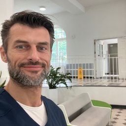 MedyFit Sp z o.o. zabawa ruchem - Uśmiechnięty mężczyzna w granatowym stroju medycznym z identyfikatorem, stojący w jasnej poczekalni z nowoczesnymi kanapami i roślinami.