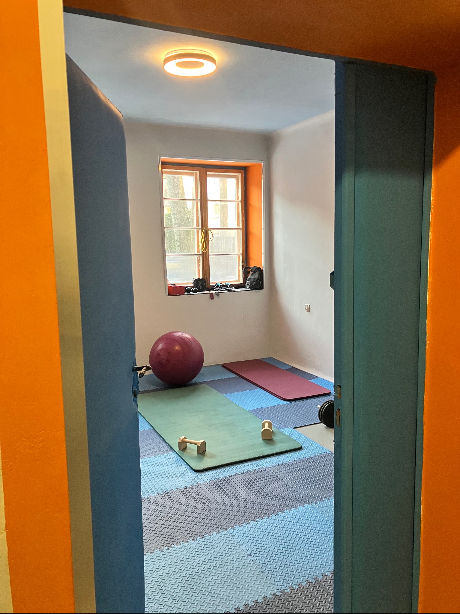 Widok zza uchylonych drzwi na salę ćwiczeń: mata, piłka, ciężarki, okno. Podłoga wyłożona puzzlami piankowymi. Pomieszczenie oświetlone lampą sufitową.