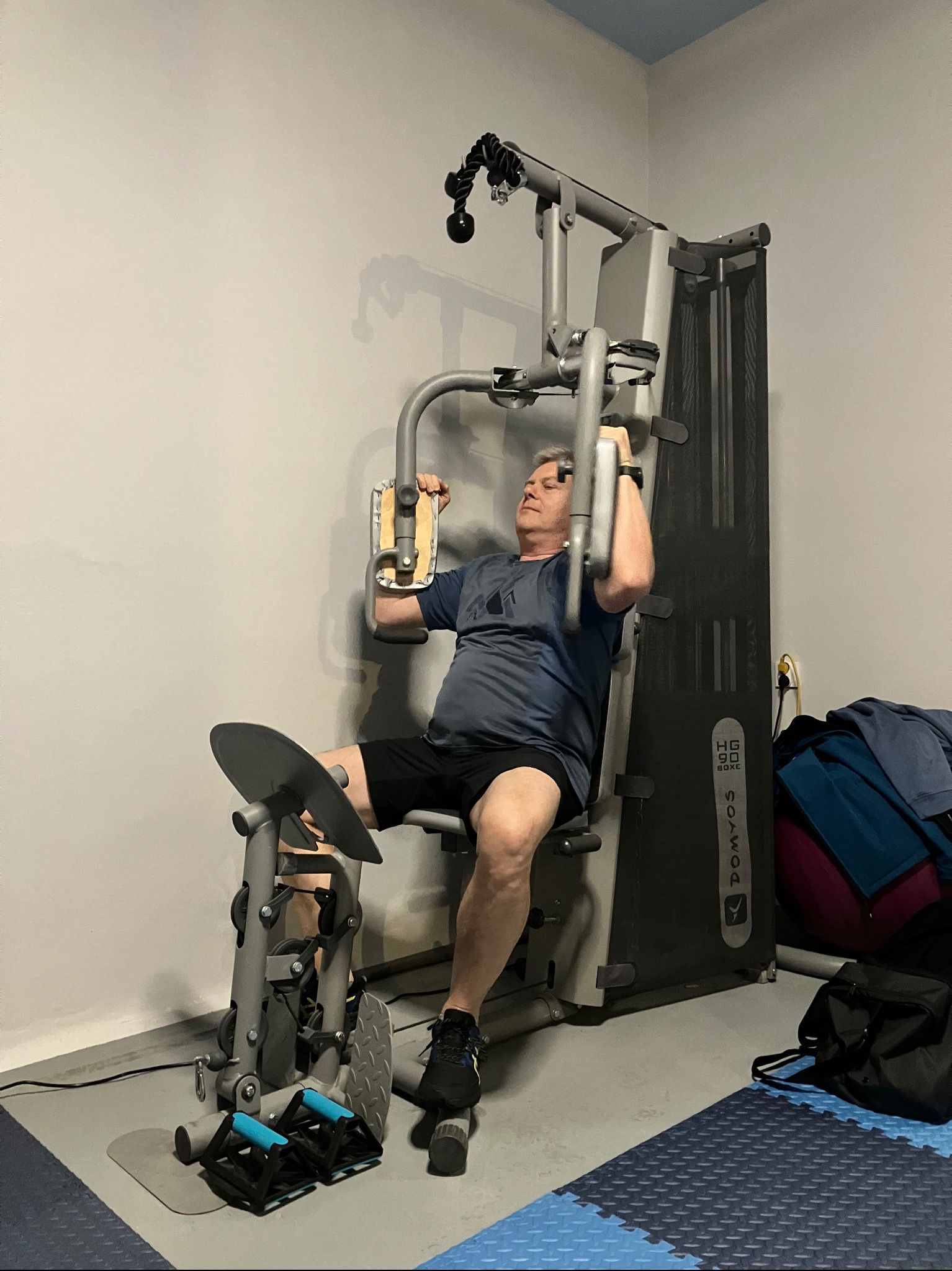 Mężczyzna ćwiczy na siłowni domowej, używając maszyny do wyciskania klatki piersiowej. Widoczne obciążenia, elementy konstrukcyjne i akcesoria do ćwiczeń.