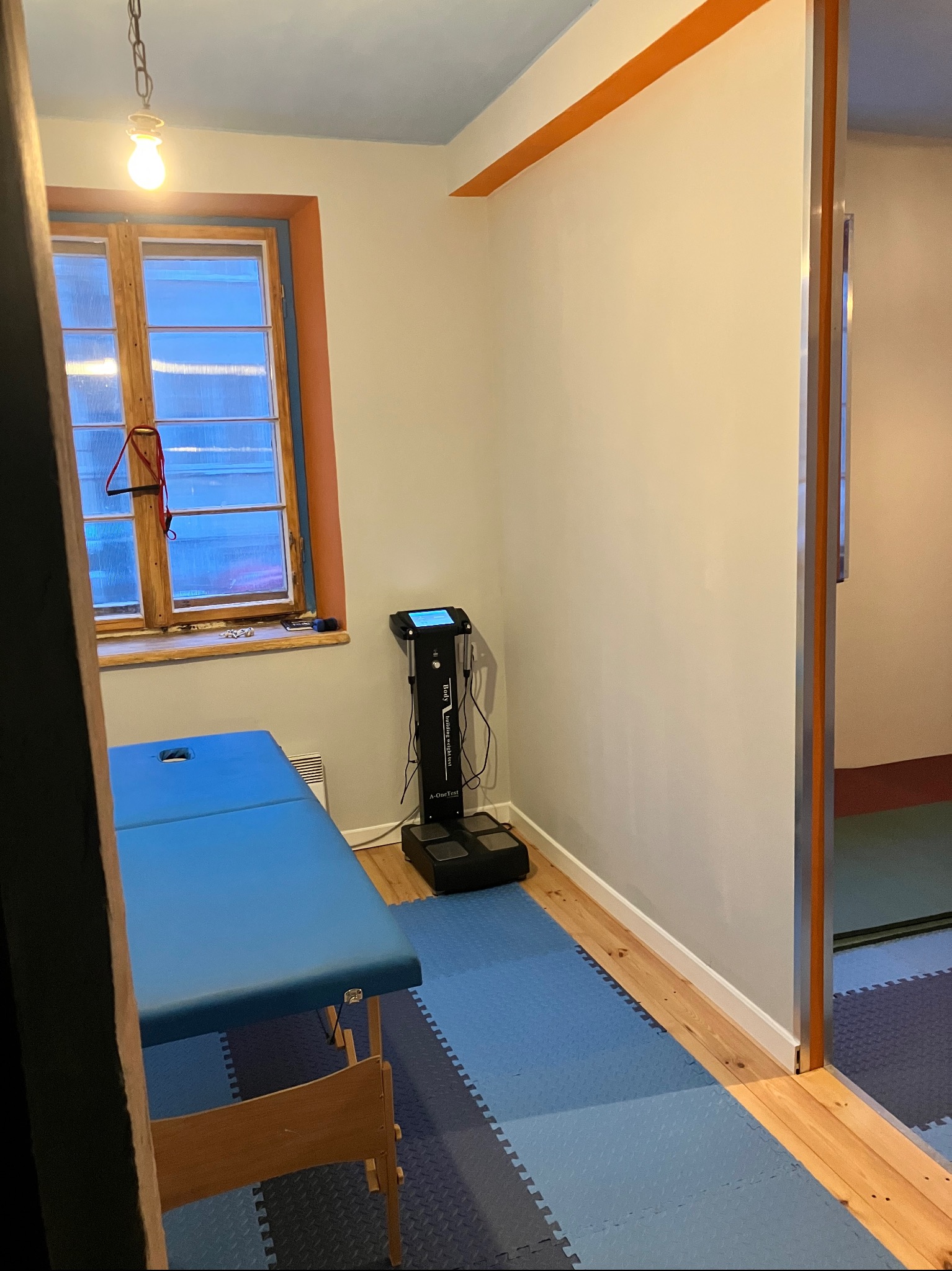 Gabinet z niebieskim łóżkiem do masażu, analizatorem składu ciała i oknem z widokiem na zewnątrz. Podłoga wyłożona niebieską pianką.