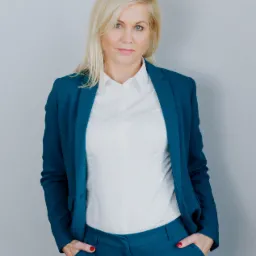 Profesjonalna blondynka w turkusowym garniturze i białej koszuli, stojąca z rękami w kieszeniach na tle szarej ściany.