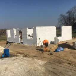 Wznoszone ściany parterowego domu z białych bloczk&oacute;w betonu kom&oacute;rkowego na fundamencie, widoczna betoniarka, taczka i sterta bloczk&oacute;w na placu budowy w słoneczny dzień.