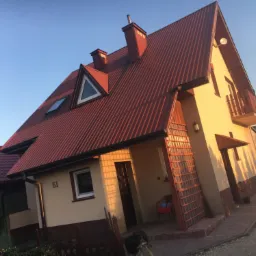 Ukośne ujęcie beżowego domu z czerwoną, skośną dachówką, widoczne okna dachowe, balkon z drewnianą balustradą oraz fragment posesji z numerem 51 i psem w kadrze.