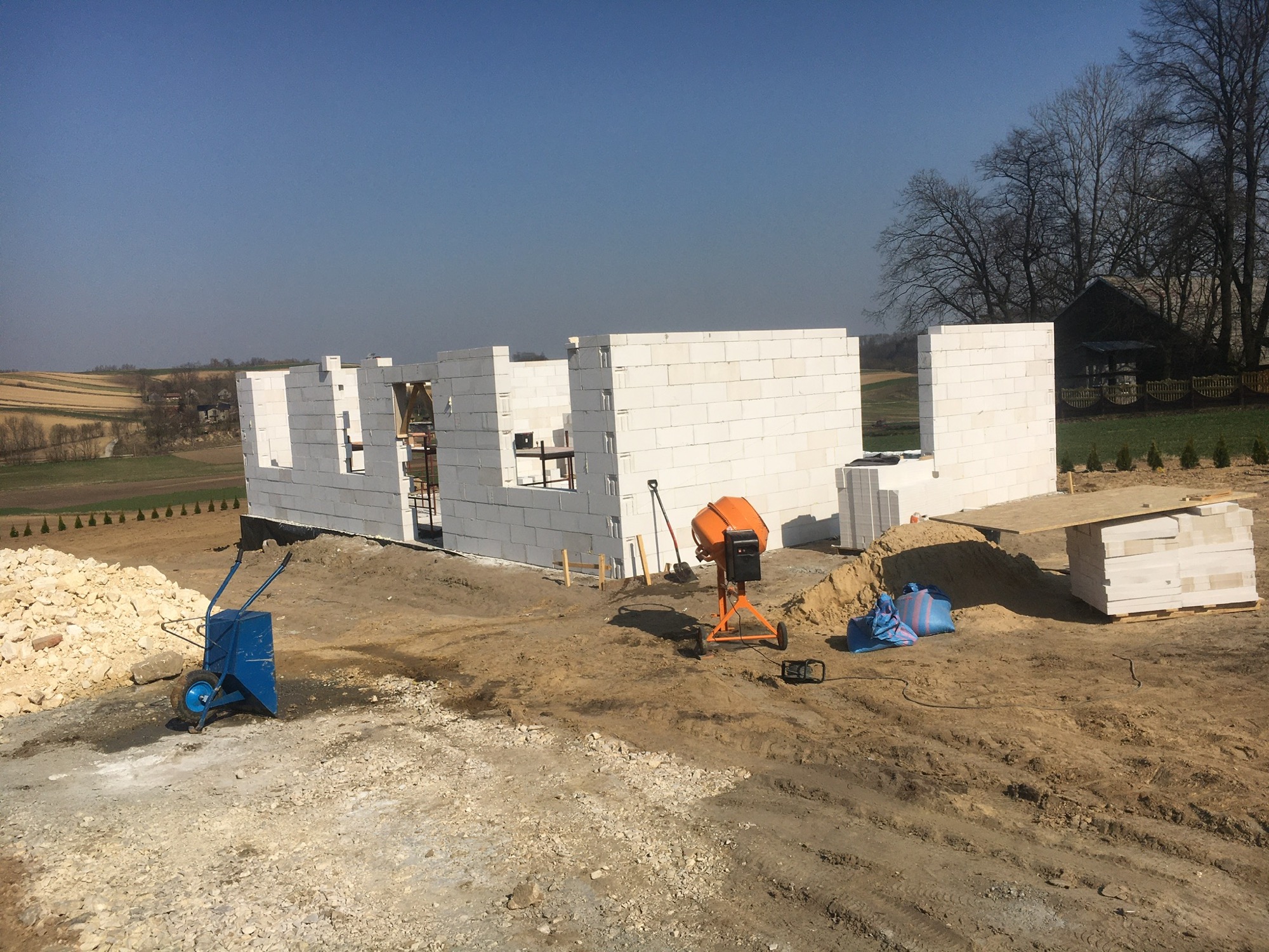 Wznoszone ściany parterowego domu z białych bloczków betonu komórkowego na fundamencie, widoczna betoniarka, taczka i sterta bloczków na placu budowy w słoneczny dzień.