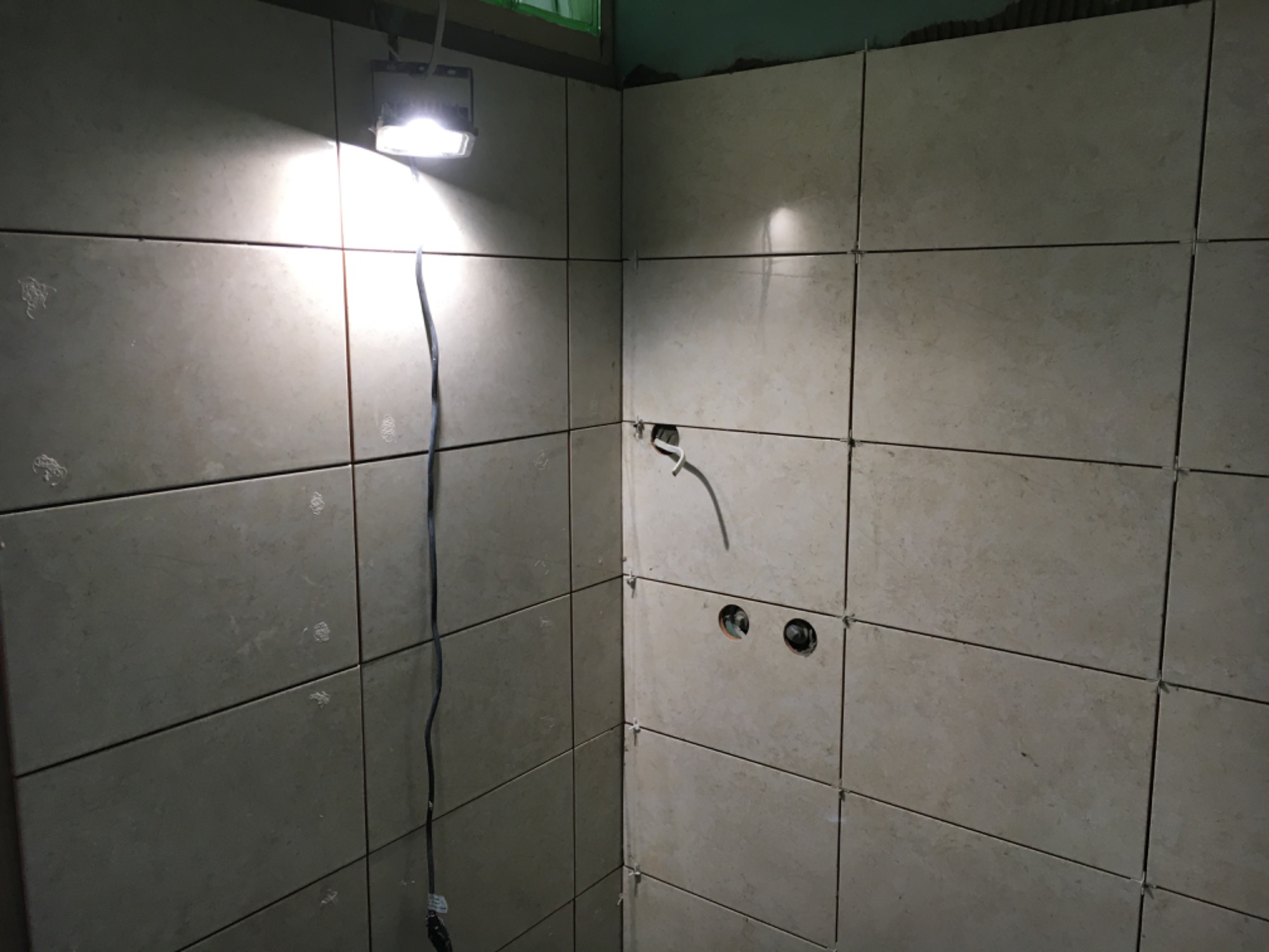 Ściany łazienki w trakcie układania płytek ceramicznych, widoczne krzyżyki dystansowe, otwory na instalacje i lampa robocza oświetlająca narożnik.