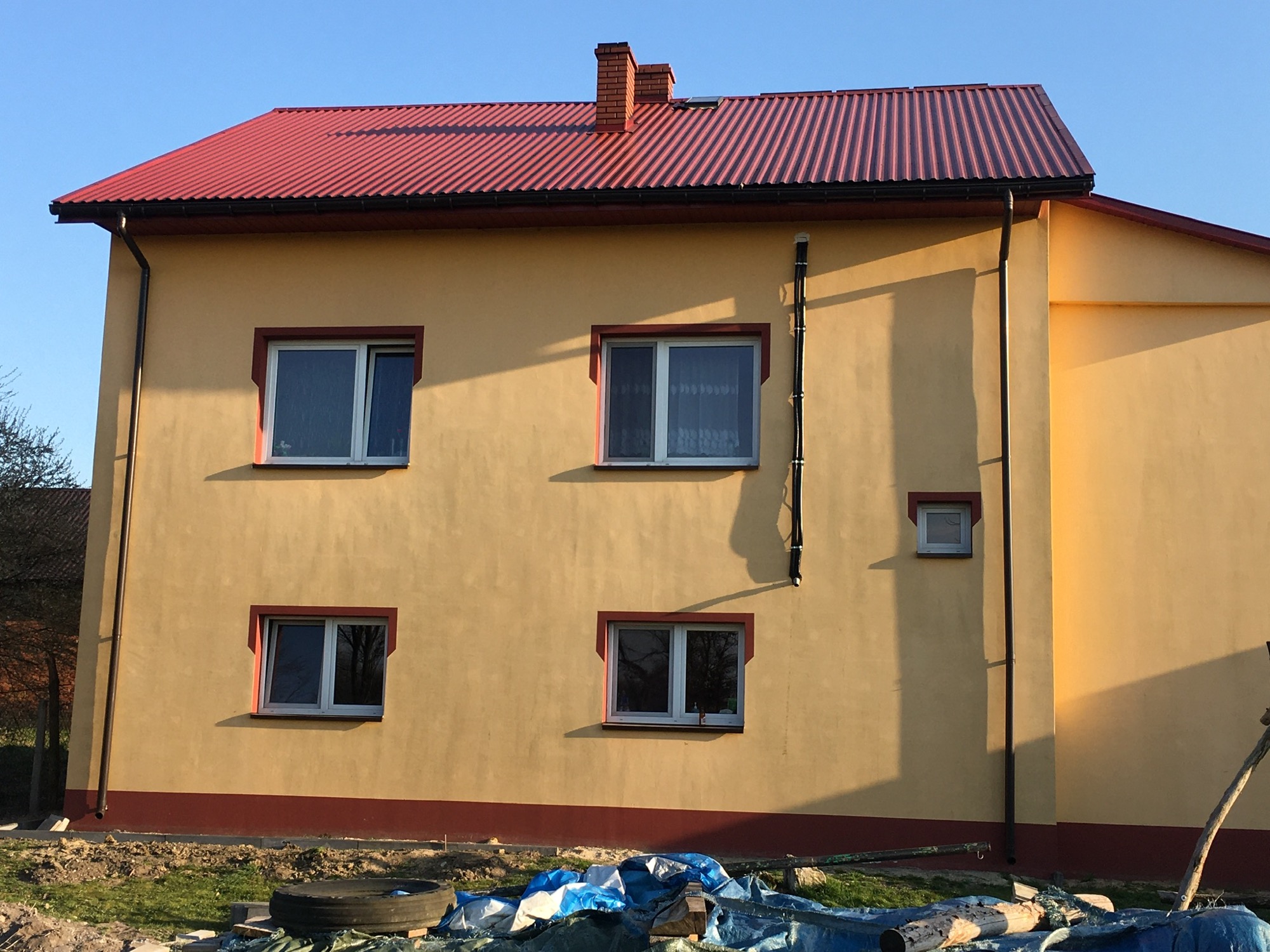 Dwukondygnacyjny dom z żółtą elewacją i czerwonym dachem z blachy trapezowej, widoczne okna z białymi ramami i czerwonym obramowaniem, rynny i pionowe rury na elewacji, teren wokół domu...