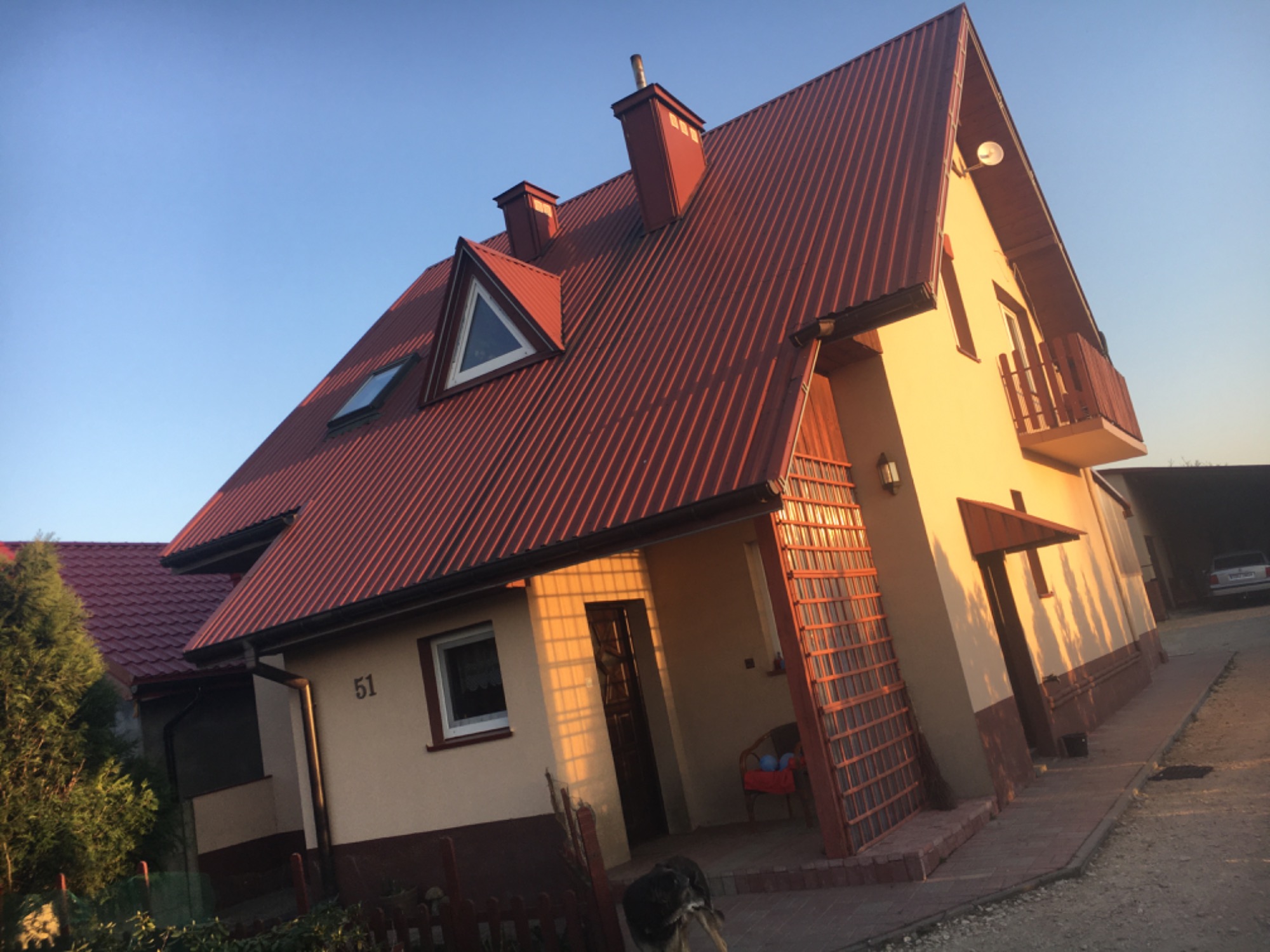 Ukośne ujęcie beżowego domu z czerwoną, skośną dachówką, widoczne okna dachowe, balkon z drewnianą balustradą oraz fragment posesji z numerem 51 i psem w kadrze.