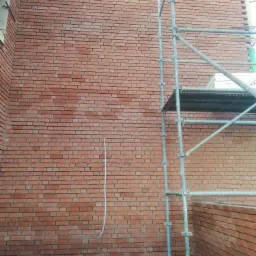 Ściana z czerwonej cegły w trakcie murowania, widoczne rusztowanie i fragment wystającego przewodu.
