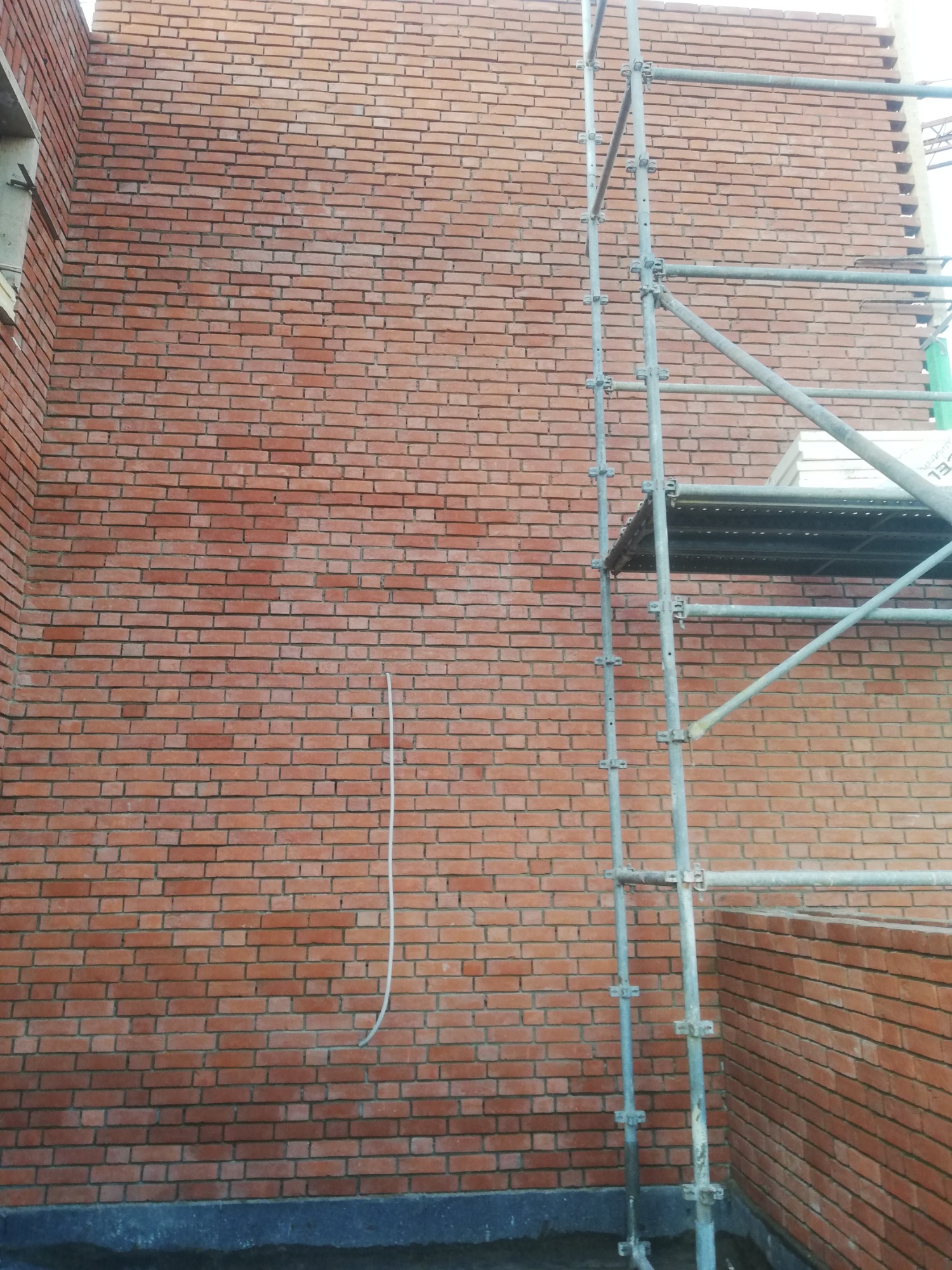 Ściana z czerwonej cegły w trakcie murowania, widoczne rusztowanie i fragment wystającego przewodu.