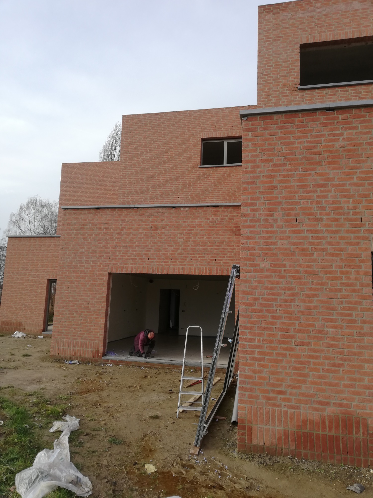 Wznoszenie surowej bryły budynku z cegły klinkierowej, widoczna drabina i pracownik przy wejściu do garażu, plac budowy z resztkami materiałów.