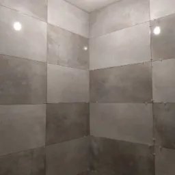 Ściany łazienki w trakcie układania płytek w odcieniach szarości, z widocznymi krzyżykami dystansowymi i fragmentem instalacji wodnej.