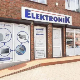 Fasada budynku firmy Elektronik z reklamami anten i dekoderów, monitoringu, podzespołów elektronicznych i serwisu RTV. Widoczne okna wystawowe i drzwi wejściowe z godzinami otwarcia.