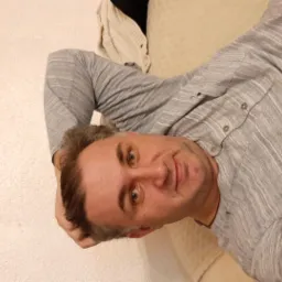 Portret mężczyzny w szarej koszuli leżącego na beżowej sofie, z widocznymi włosami i fragmentem kremowego swetra w tle.